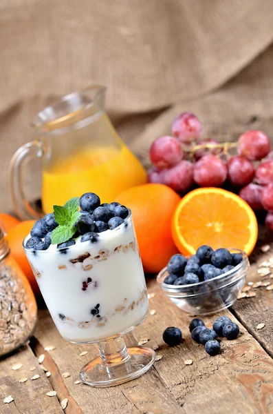 Griechischer Joghurt mit Haferflocken, frischen Blaubeeren und Minzblättern sowie Orangen und Trauben im Hintergrund — Stockfoto