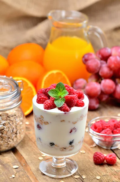 Griechischer Joghurt, frische Himbeeren im Glas und Minzblätter, Orangen, Trauben und ein volles Glas Haferflocken im Hintergrund - gesundes Frühstückskonzept — Stockfoto