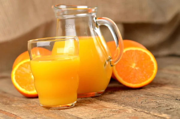 Vaso lleno de zumo de naranja, jarra con zumo fresco y naranjas frescas en el fondo sobre mesa de madera — Foto de Stock