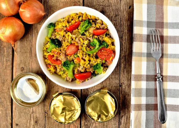 Blick von oben auf Schüssel mit Thunfisch-Risotto mit Gemüse, Tomaten, Brokkoli und Petersilie, Zwiebeln, Dosen und braun karierter Tischdecke im rechten Rahmen — Stockfoto