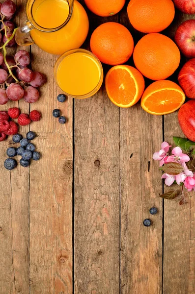 Sumo de laranja, laranjas frescas, maçãs, uvas, framboesas, mirtilos e flores de primavera em uma mesa de madeira - fundo de fruto - visão de cima - foto vertical — Fotografia de Stock