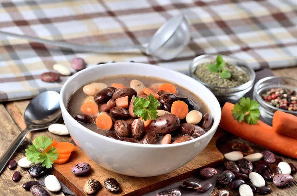 Schüssel Bohnensuppe mit großen Bohnen auf Schneidebrett, Karotten, Petersilie, Majoran, Löffel, Handtuch und Schöpfkelle im Hintergrund — Stockfoto