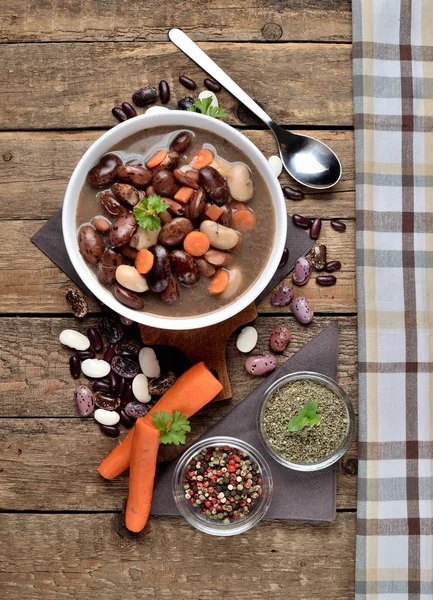 Nahaufnahme einer vollen Schüssel heiße Bohnensuppe mit großen Bohnen auf Schneidebrett, Karotten, Petersilie, Majoran, Löffel, Handtuch und Schöpfkelle im Hintergrund - von oben, vertikales Foto — Stockfoto