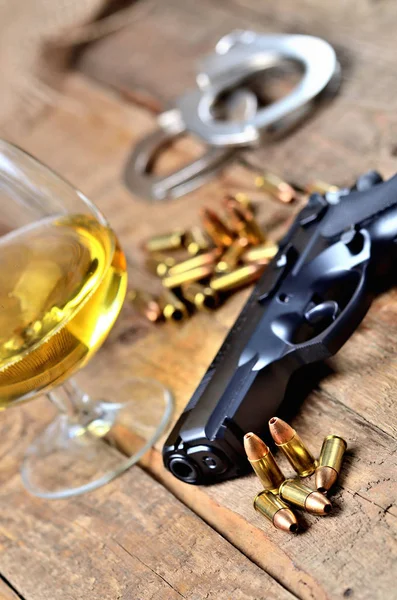 Ποτήρι κονιάκ, πιστόλι 9mm, χειροπέδες και σφαίρες στο παλιό ξύλινο τραπέζι - κάθετη φωτογραφία — Φωτογραφία Αρχείου