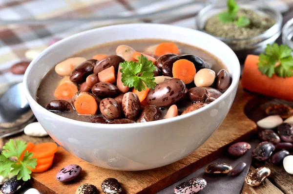 Schüssel Bohnensuppe mit großen Bohnen auf Schneidebrett, Karotten, Petersilie, Majoran, Löffel und Schöpfkelle, Handtuch im Hintergrund — Stockfoto