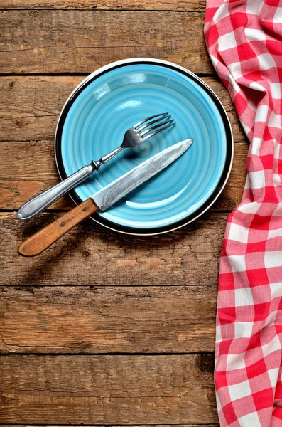 Mavi seramik tabak ve kırmızı kareli masa örtüsü sol çerçeve üzerinde eski vintage ahşap masa arka plan - yukarıdan görüntüleyin bıçak ve çatal — Stok fotoğraf