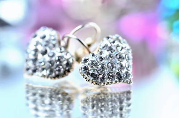 Close-up of pair of heart shaped earring decorated with diamonds on glass and elegant reflection - colored background blue and purple — Stock Photo, Image