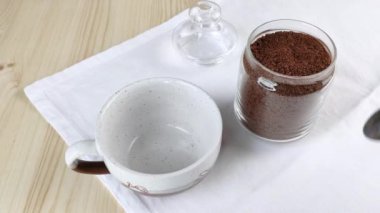 Cam kavanozdan dondurulmuş kahve kaşıkla bardağa konur. Kafein içeceği yapıyorum. Yakın plan, tahta masa, beyaz peçete.