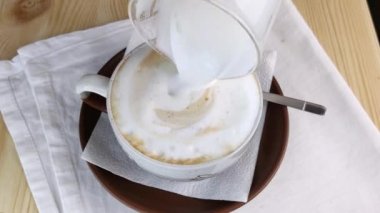 Kapuçino ya da latte. Köpük sütü yuvarlak kahve bardağına dökülür. Kremalı köpükle içki yapıyorum. Yakın çekim, beyaz peçete, ahşap masa.