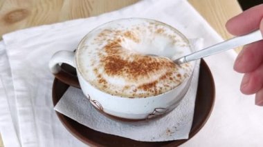 Kapuçino ya da latte. Kaşık bir fincan kahveden süt köpüğünü toplar. Köpük üzerine tarçın ve kakao tozu serpilmiş. Kremalı içecek yapıyorum. Yakın plan, beyaz peçete, tahta masa.