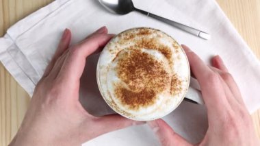 Kapuçino ya da latte. İki el, üzerinde tarçın ve kakao tozu serpiştirilmiş süt köpüğü olan bardağı tutar. Kremalı köpükle iç. Yakın çekim, beyaz peçete, ahşap masa.