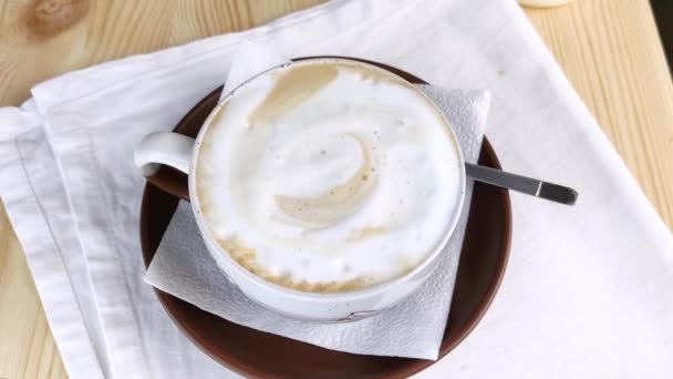 卡布奇诺或拿铁咖啡 咖啡与泡沫牛奶在圆杯与褐色茶托和勺子是在桌子上 用奶油泡沫喝 特写顶景 白色餐巾 木制背景 — 图库视频影像