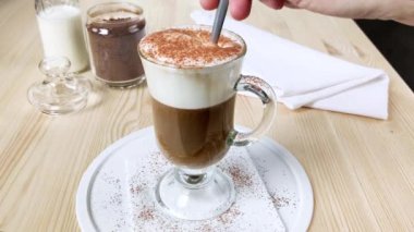 Kapuçino, fincanda köpüklü süt olan latte. El, kaşık dolusu kahveyi uzun bardakta karıştırır, kremayla karıştırır, tarçın ve kakao tozu serpiştirir. İki katlı tatlı içeceği. Yakın kenar görünümü