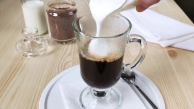 Kapuçino ya da latte. Köpük sütü bir bardak kahveye dökülür. Uzun, berrak bardakta, saplı kremalı köpükle içki yapmak. Yakın plan, beyaz tepsi, ahşap masa, kavanozda granüle kahve.