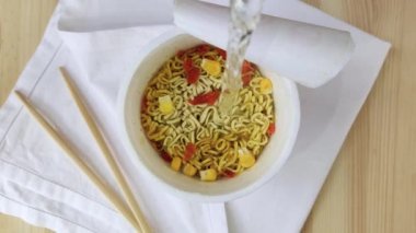 Kadehteki yumurtalı erişteler kaynar suyla doldurulur. Anında ramen çorbası. Tavuk aromalı fast food, kurutulmuş havuç, mısır. Yakın çekim üst manzara, ahşap masa, beyaz peçete, çubuklar, 4K