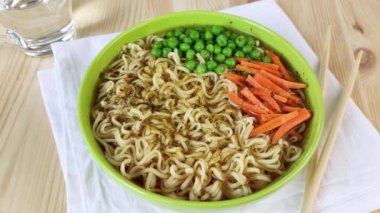 Yeşil tabakta erişte çorbası, taze bezelye ve havuç. Çiğ yumurta, erişte, et suyu ve sebzelerle dolu kaseye düşer. Evde doyurucu bir öğle yemeği pişiriyorum. Yakın plan, ahşap masa, beyaz peçete, çubuklar, 4k.