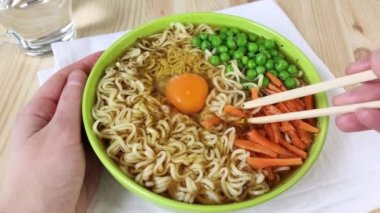 Yeşil tabakta erişte çorbası taze bezelye, havuç, çiğ yumurta. Yemek çubukları erişte, et suyu ve sebzelerle dolu kaseden yiyecek alır. Evde doyurucu bir öğle yemeği pişiriyorum. Yakın plan, ahşap masa, beyaz peçete, 4K