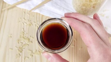 Şeffaf cam sos kabında soya sosu. Kadın el ele tutuşup yuvarlak kaseyi gösteriyor. Geleneksel Asya mutfağı malzemesi. Tahta masa, yakın plan, dağınık pirinç taneleri, yemek çubukları, beyaz peçete.