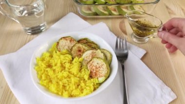 Sarı körili pilav ve fırında kabak provenkal otlarla yağlanır. Kadın eli cam sos teknesini tutuyor. Sağlıklı yemek, akşam yemeği için baharatlı sebze pişirmek. Yakın plan, beyaz peçete, tahta masa.