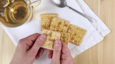 Kadın elleri etsiz kraker tutar, kuru kurabiyeleri ikiye böler. Bardakta şeffaf çay bardağı. Sağlıklı hafif atıştırmalıklar. Yakın çekim, ahşap masa, beyaz peçete.