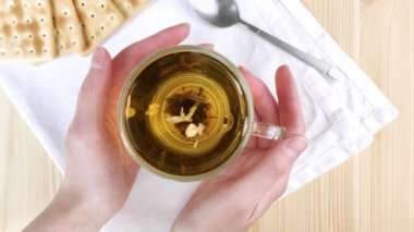 Yaseminli yeşil çay demlenir, yapraklar daireler çizer. Kadın eli, cam şeffaf bardakta sıcak içeceği tutar. Sağlıklı hafif kahvaltı. Yakın çekim üst manzara, ahşap masa, beyaz peçete, kuru kurabiye.