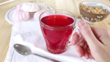 Elleri bir fincan meyve çayını tutar ve masanın üzerinde döndürür. Cam bardakta Hibiscus. Parlak kırmızı içecek, komposto. Şeker ve böğürtlenli yaz tatili. Yan görüş, beyaz peçete, lokum, frenk üzümü.