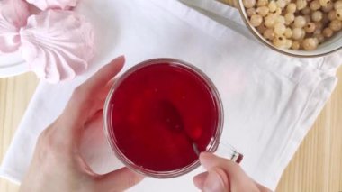 Elleri fincanı tutuyor ve bir kaşık meyve çayı karıştırıyor, üzümlü üzümleri gösteriyor. Cam bardakta Hibiscus. Parlak kırmızı içecek, komposto. Şekerli çay molası. Yakın çekim üst manzara, beyaz peçete, lokum.