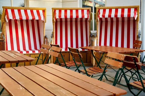 View to a group of wooden patio furniture on the terrace of a restaurant with three beach chairs in the background.