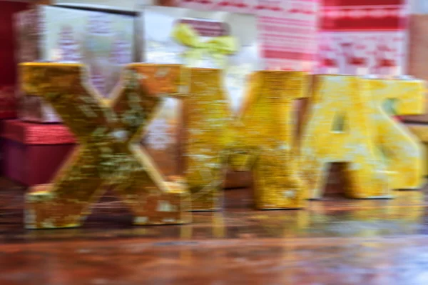 Large golden letters showing the text XMAS in front of presents and overlaid with a motion blur to serve as an abstract background.