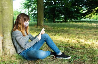 Coronavirus salgını sırasında yeniden kullanılabilir maske takıp parkta kitap okuyan çekici genç bir kadın.