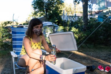 Yüz maskeli çekici genç bir kadın Coronavirus salgını sırasında kamp yaparken taşınabilir bir buzdolabından bir şişe su alıyor.