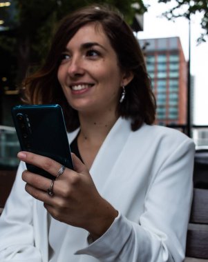 İngiltere 'de Londra' da bir bankta otururken beyaz takım elbise giyen çekici bir kadın tarafından kullanılan mavi bir akıllı telefonun yakın plan fotoğrafı.