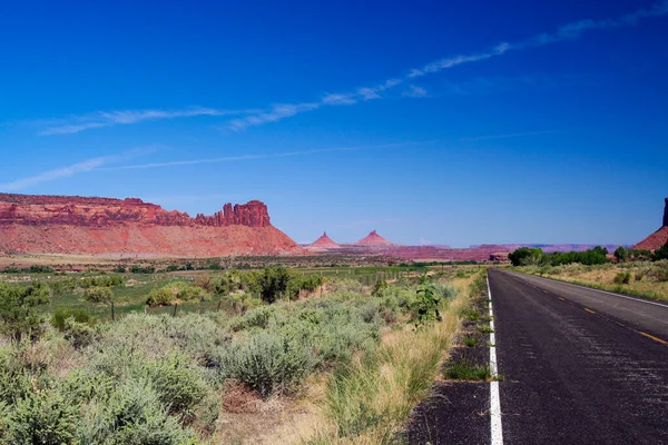 View over green valley on endless straight road with steep red sandstone hill formation (butte) against blue sky  - Monument Valley, Utah, Arizona