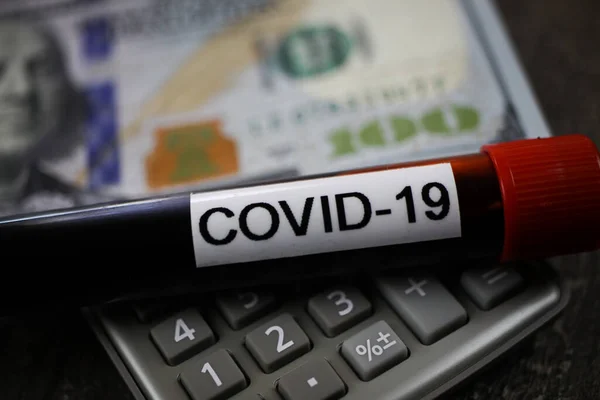 Calculation of economic damage concept: covid-19 blood sample tube on calculator, blurred 100 us dollar bill background (focus on keys lower third)