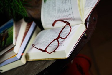 VİERSEN, ALMANY - 11 Mart. 2019: Kırmızı okuma gözlüklü ve gece vakti antika yuvarlak ahşap masadaki seyahat kitapları yığınıyla açık bir kitap görüntüsü. Sulu yeşil bitki arkaplanı.