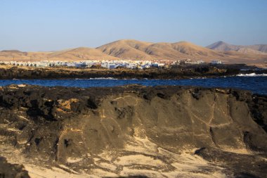 El Cotillo: Kuzey Fuerteventura arka planında kuru kahverengi tepeli beyaz köydeki volkanik kayalar ve mavi lagünün üzerine bakın