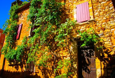Akdeniz 'de pembe pencere panjurları, ahşap kapı ve asma bitkisi olan taştan ev duvarına bakın - Roussillon en Provence, Fransa