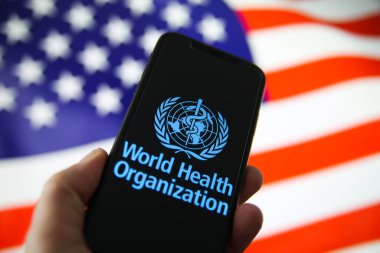 Viersen, Almanya - 9 Temmuz. 2020: Dünya Sağlık Örgütü (WHO) logosu cep telefonu ekranında insan eliyle tutuluyor. ABD arkaplanının bulanık bayrağı. (Logoya odaklan)