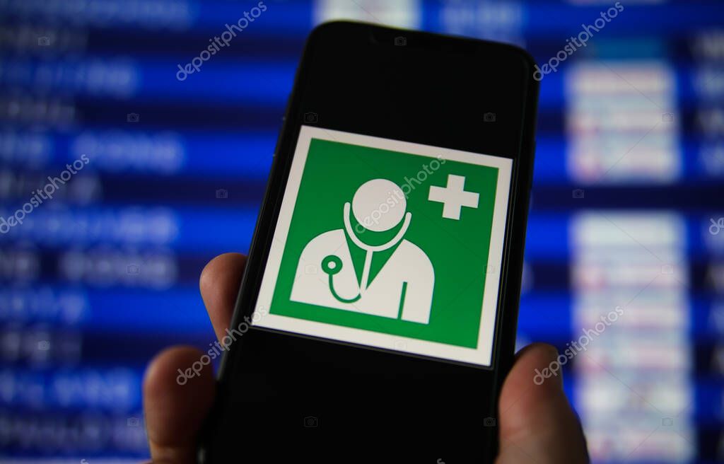 Viersen, Germany - July 9. 2020: View on hand holding mobile phone with green sign for health and medical assistance at airport. Blurred departure board background.