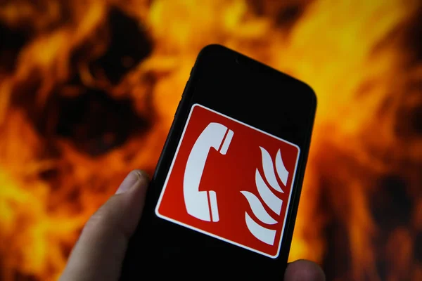 View on hand holding mobile phone with international fire emergency telephone symbol. Blurred fire flames background. (Selective focus on upper phone receiver)