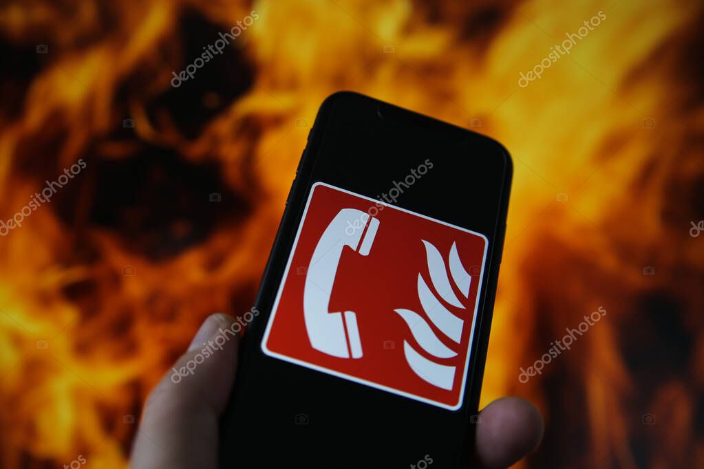 View on hand holding mobile phone with international fire emergency telephone symbol. Blurred fire flames background. (Selective focus on upper phone receiver)