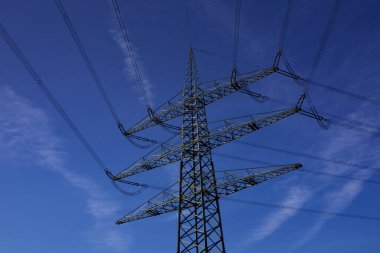 Bir elektrik direği, Almanya 'da güneş parlarken az bulutlu mavi bir gökyüzünün önünde birçok kabloyla durur.