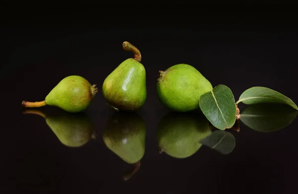 Three small green pears and a leaf lie against a dark background and are reflected