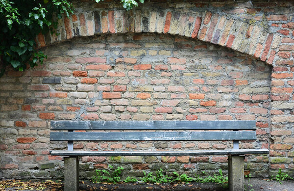 An old wooden bench stands in front of a historic wall made of burnt red bricks with a round arch and ivy