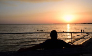 Bir adam önde oturur ve sırtını fotoğrafçıya çevirir ve kollarını uzatarak güneşin akşamları battığı denizin ufkuna bakar.