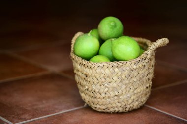 Küçük bir sepetin içinde yeşil organik limonlar var. Kahverengi fayansların üzerinde koyu bir arka plan var.