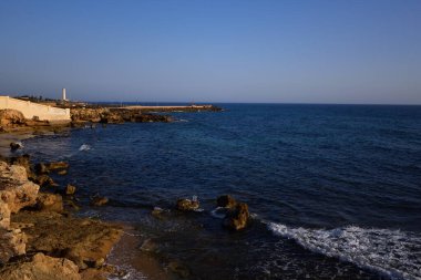 Sahili, kayaları ve arka planda deniz feneri olan denizin manzarası, İtalya 'da mavi bir gökyüzüne karşı.