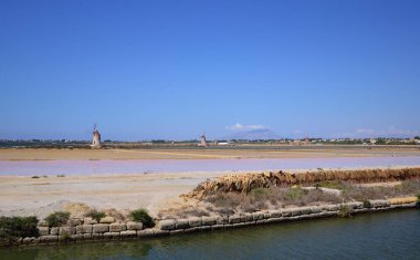 Sicilya 'da Marsala yakınlarında deniz kenarındaki tuz panorama, kırmızı tuz havzaları ve yel değirmenleriyle, mavi gökyüzüne karşı, bir kanalın önünde