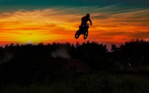 Motocross pilot in a jump during sunset with smoke on dirt track.