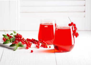 İki bardak soğuk kırmızı frenk üzümü suyu beyaz ahşap panjurların arka planında duruyor, yanında da naneli, yatay oryantasyonlu frenk üzümü var..